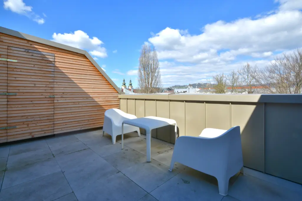 Moderne Dachterrasse mit zwei weißen Sesseln, Tisch und Ausblick auf Stadt und Hügel.